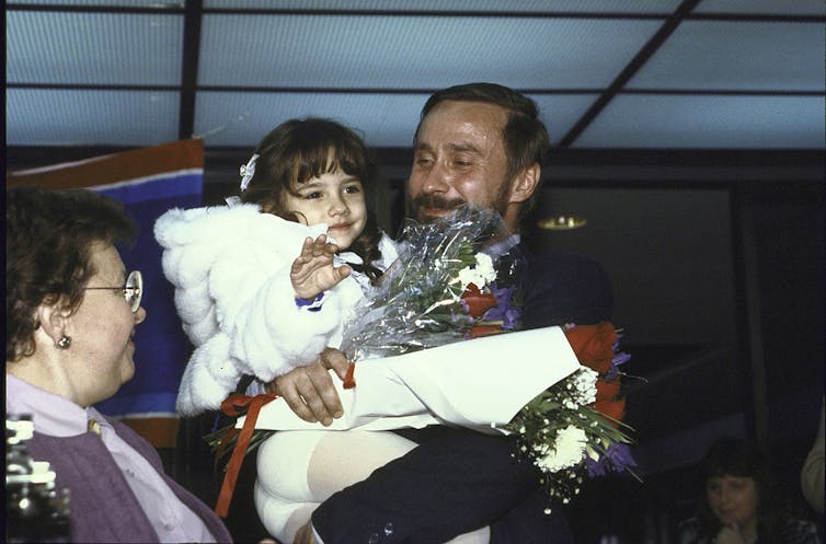 A man in a suit smiles and holds a young girl in a white jacket, who waves at the camera.