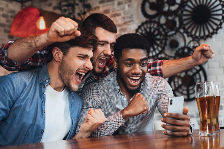 Three men cheer while gathered around a mobile phone, they are sitting at a bar with a pint of beer