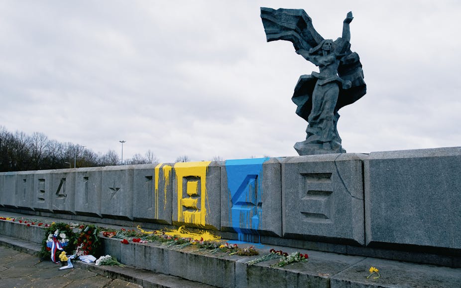 Statue of a woman on a wall with war dates, splashed with yellow and blue paint