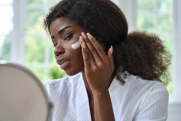 A young woman applies sunscreen to her cheek indoors.
