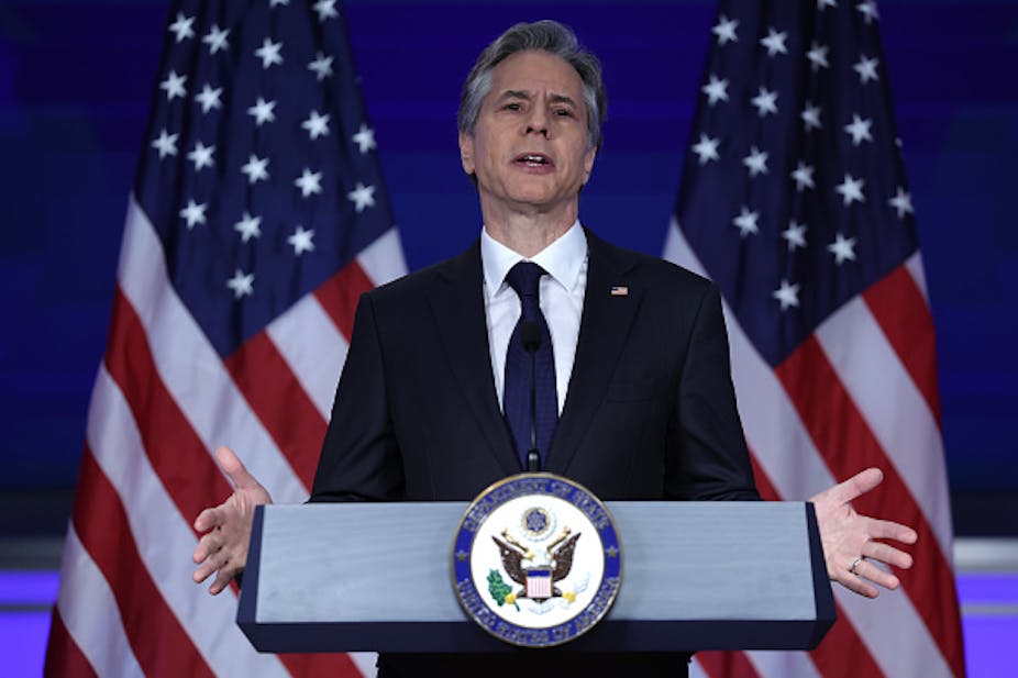 A man gestures with both arms while speaking at a podium with his back to two American flags.