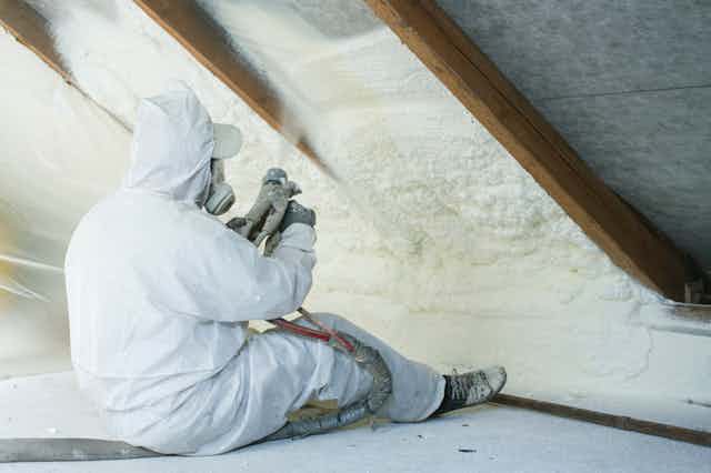 A worker in white overalls sprays white foam into the eaves of a house.