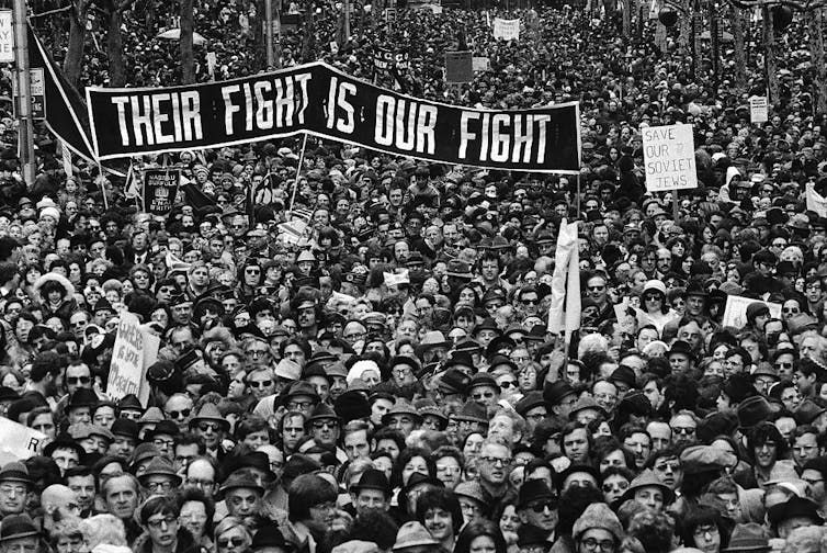 A black and white photo shows a closely packed crowd at a protest, with a large sign that says 'Their fight is our fight.'