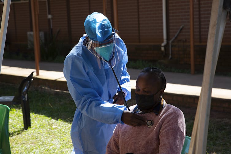 A health-care worker in blue protective garb listens to the breathing of a woman wearing a mask and a pink shirt.
