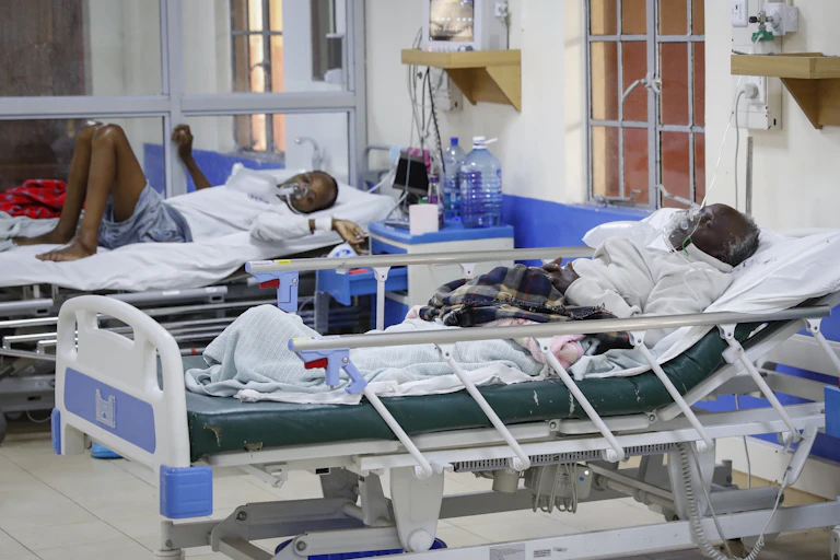 Two people lie in separate hospital beds, one young and one aged. Both wear oxygen masks.