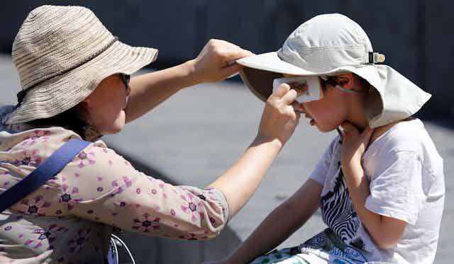 A woman wearing a beige hat and sunglasses applies sunscreen to a child earing a white hat