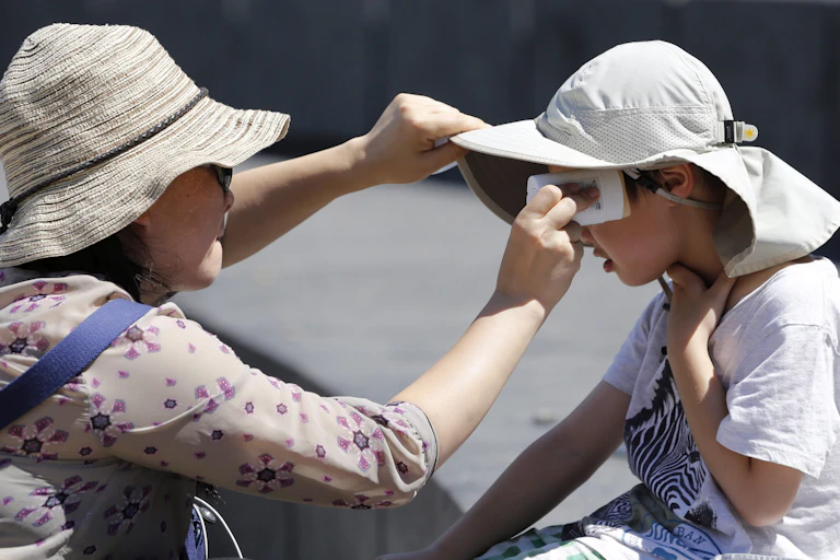 A woman wearing a beige hat and sunglasses applies sunscreen to a child earing a white hat