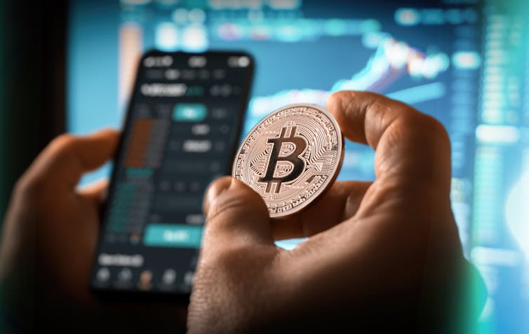 persona sostiene una moneda Bitcoin frente a una pantalla