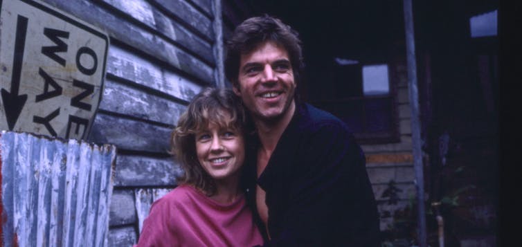 a woman and man, smiling, stand in front of a weathered wall, the side of a house
