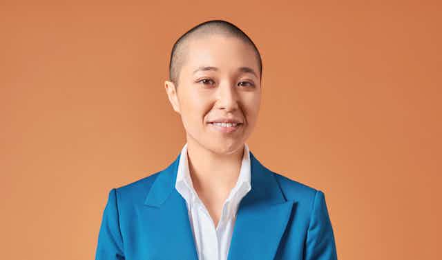 a smiling nonbinary person with a shaved head, in a blue suit and white shirt