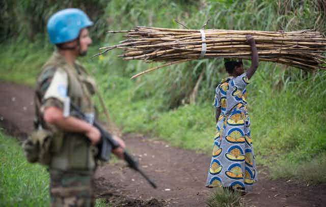Soldier with blue helmet on patrol as villager gathers wood.