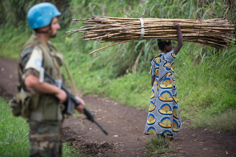 Soldier with blue helmet on patrol as villager gathers wood.