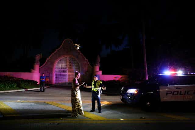 A woman in a long dress talks to a police officer outside a very fancy gate, in the night.