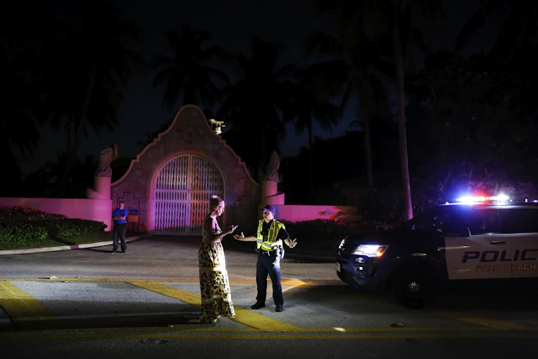 A woman in a long dress talks to a police officer outside a very fancy gate, in the night.