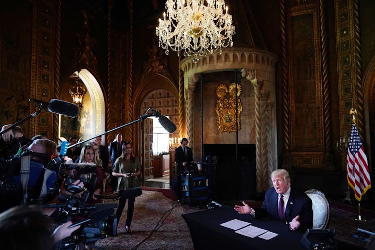An older white man is shown seated at a desk, gesticulating with his mouth open, in an ornate-looking room. In front of him is a group of reporters and camera people with equipment.