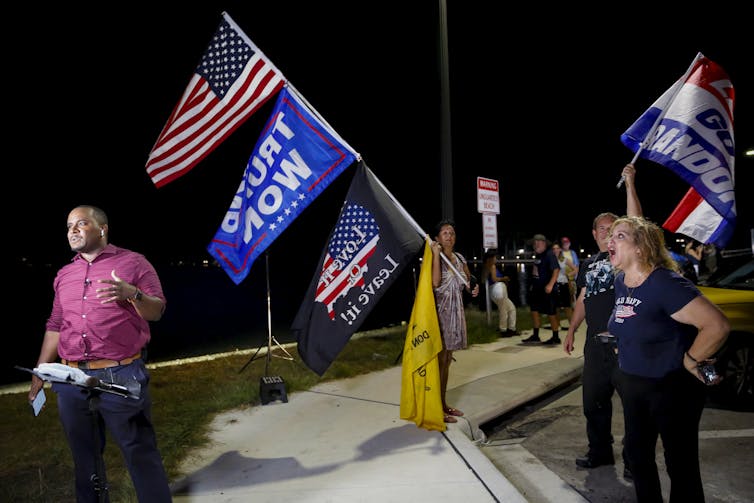 A few people - one of them yelling - are shown with Trump flags and American flags on a dark evening on the street.