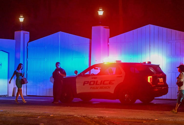 A police officer leans against a police car while a woman walks past. Behind them are large white gates, shining blue and red because of the police lights.
