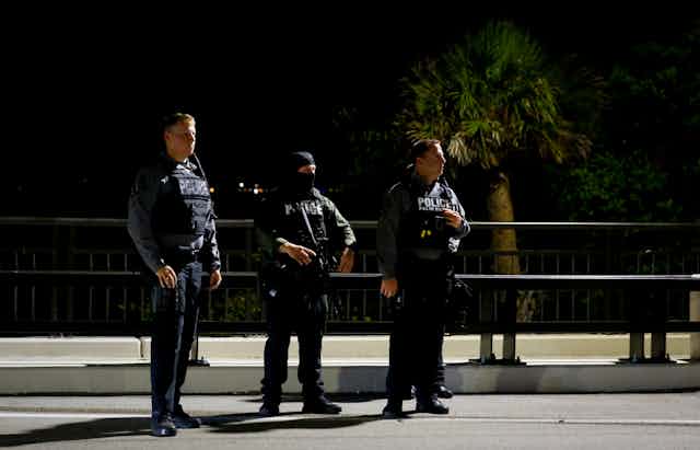 Three male police officers in tactical vests stand on a quiet street at night with palm trees behind them.