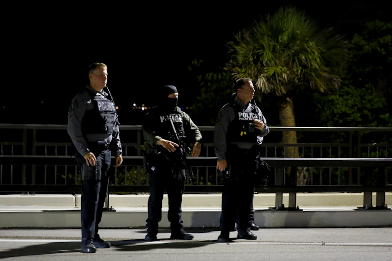 Three male police officers in tactical vests stand on a quiet street at night with palm trees behind them.