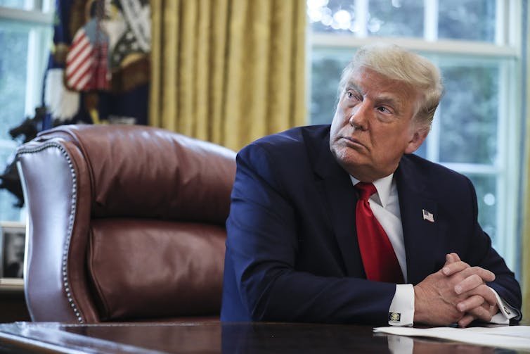 A middle-aged white man is dressed in a navy blue business suit and sitting on a leather chair behind a wooden desk.