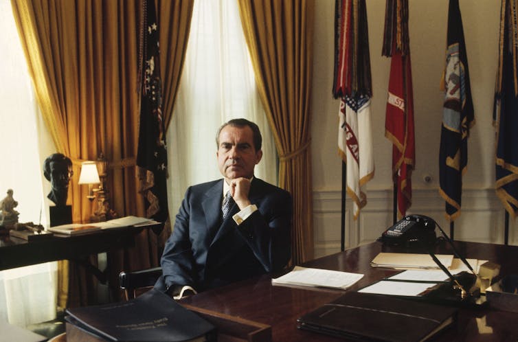 A middle-aged white man sits behind a desk and poses for a portrait.