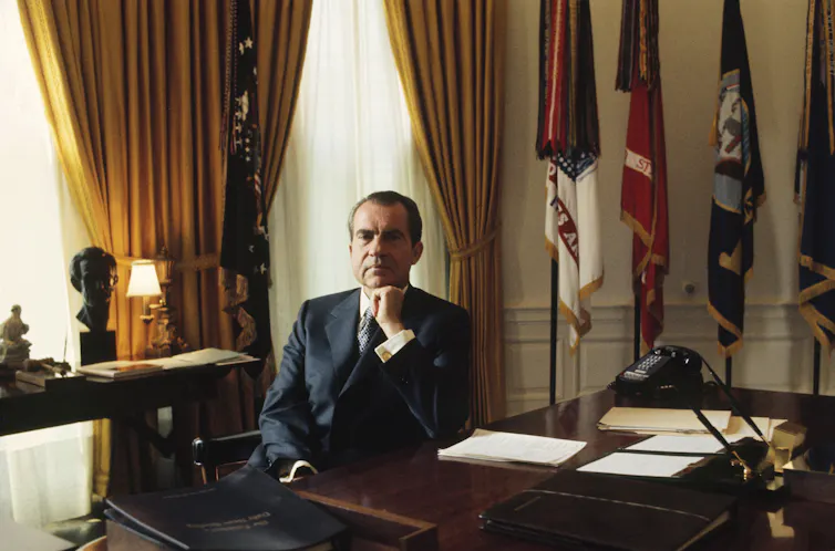 A middle-aged white man sits behind a desk and poses for a portrait.