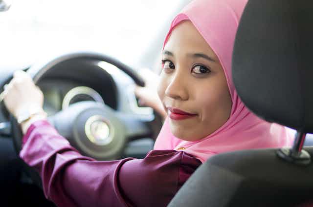 A smiling woman in a pink hijab sits at the wheel of a car and looks over her shoulder