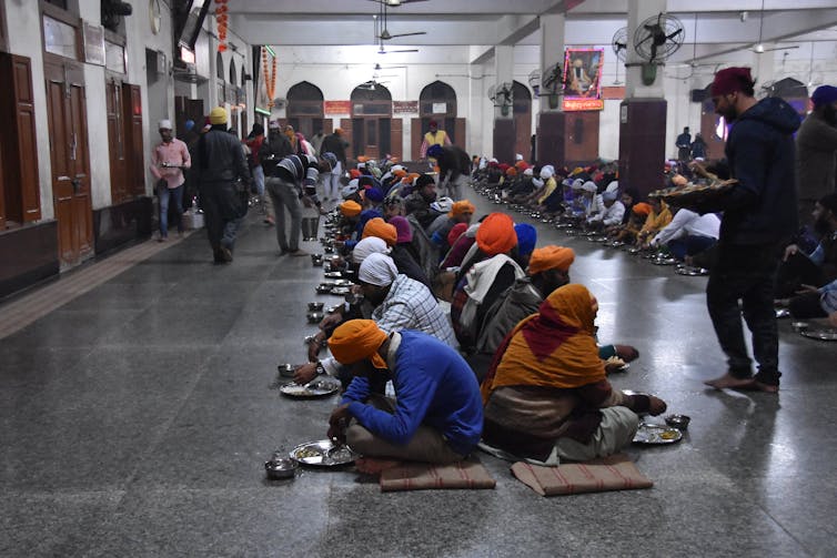 Men and women seated on the floor in two rows, being served food.