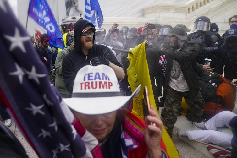 A woman wearing a 'team trump' cowboy hat carries an american flag. Behind her rioters confront police wearing riot gear.