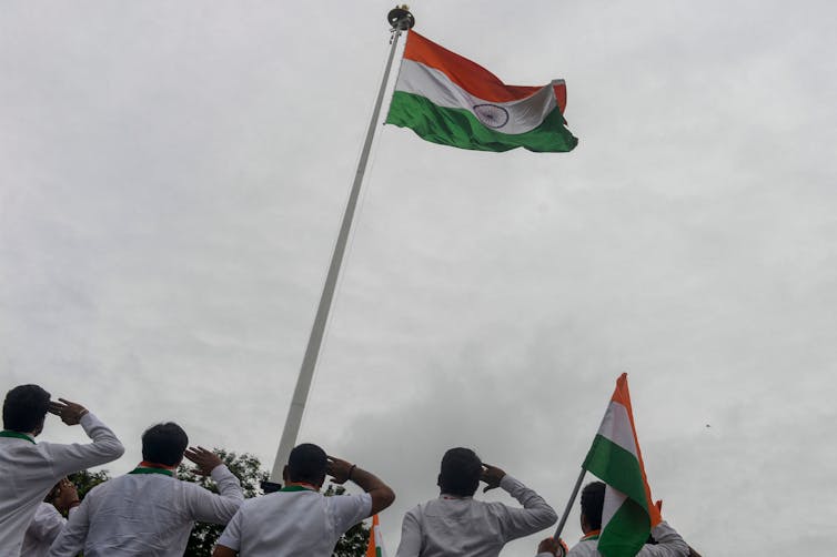 Five men in bottom of frame look up at an Indian tricolor flag flying high, while saluting