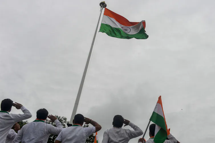 Five men in bottom of frame look up at an Indian tricolor flag flying high, while saluting