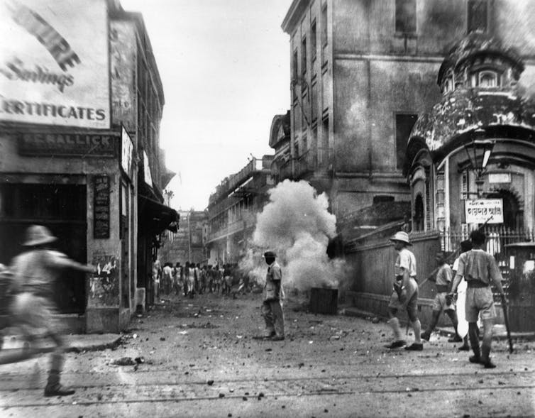Black-and-white photo of a city street with police running and tear gas explosions in the background