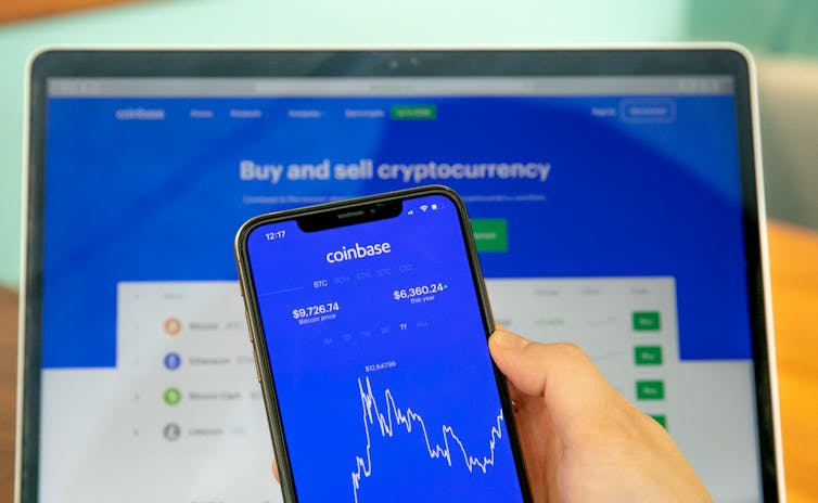 A hand holding a cellphone with the Coinbase app open on it in front of a laptop with the Coinbase website open