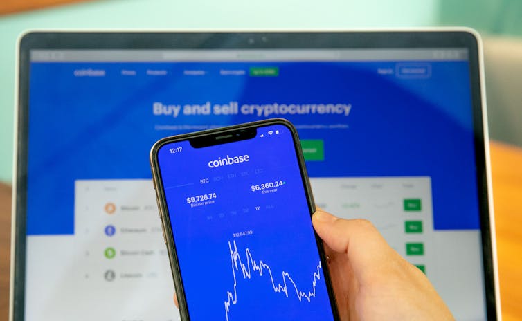 A hand holding a mobile phone with the Coinbase app open in front of a laptop with the Coinbase website open