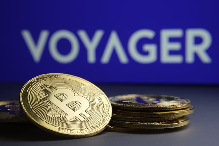 A stack of bitcoins sit in front of the logo for cyptocurrency company Voyager