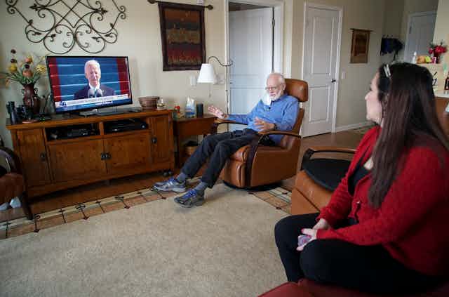 a man and a woman sit in a living room while a television in the background displays President Biden speaking at a podium