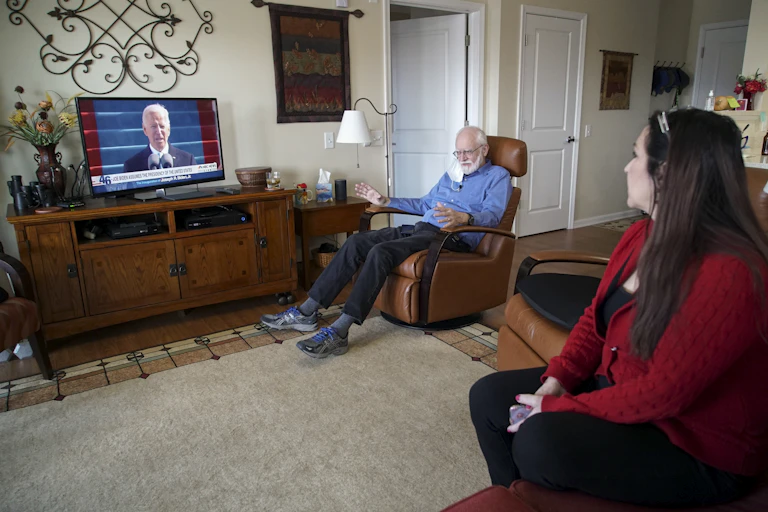 a man and a woman sit in a living room while a television in the background displays President Biden speaking at a podium