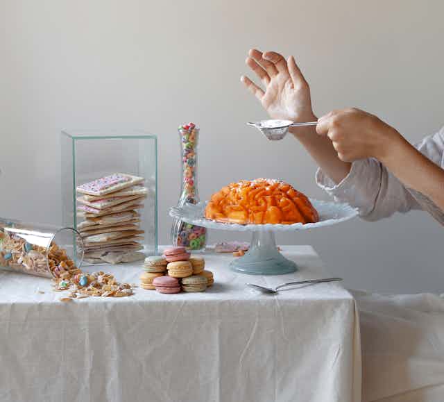 A jello mold in the shape of a brain sits on a platter, surrounded by sweets, while a person in the background holds a spoonful of sugar above the mold.