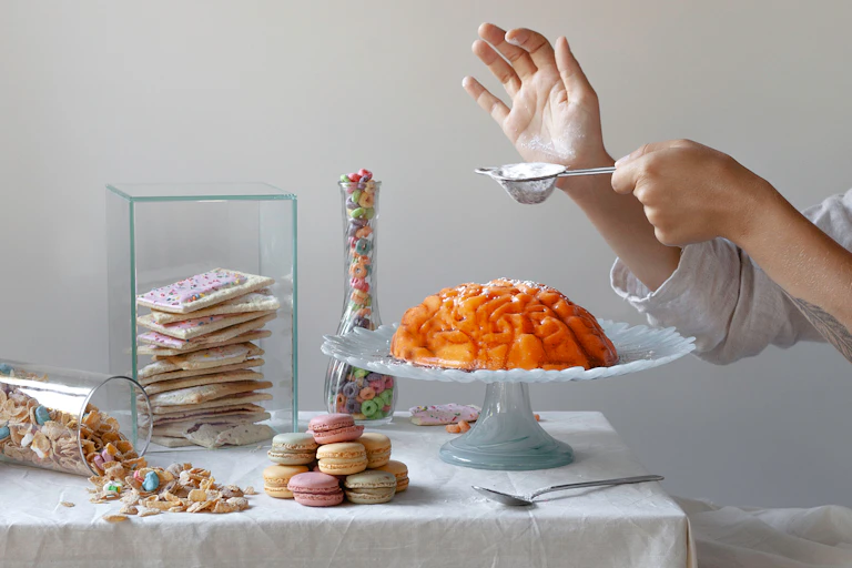 A jello mold in the shape of a brain sits on a platter, surrounded by sweets, while a person in the background holds a spoonful of sugar above the mold.