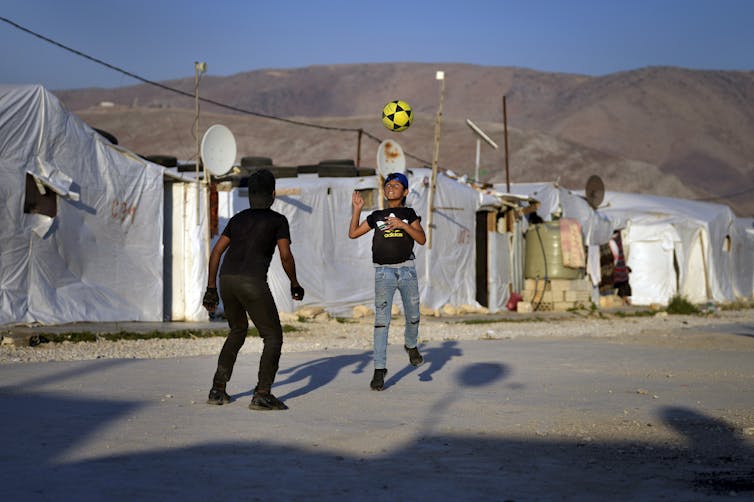 Two boys play soccer with a black and yellow soccer ball with tents and brown hills in the background.