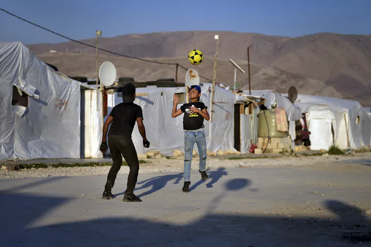 Two boys play soccer with a black and yellow soccer ball with tents and brown hills in the background.