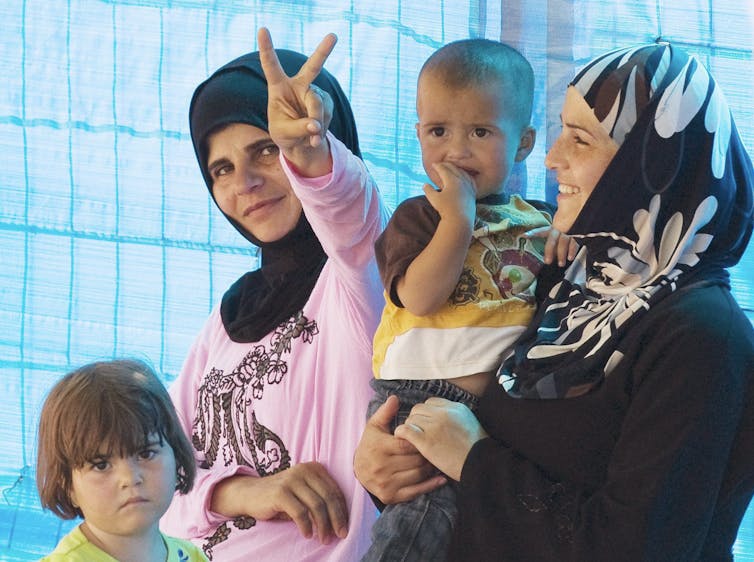 Two women in head scarves smile. One holds a baby while another flashes a V for victory sign.