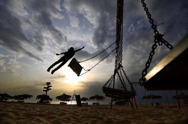 A boy is seen in silhouette jumping off a swing at a beach.