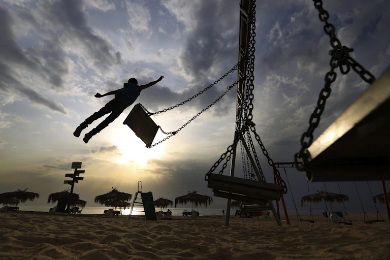 A boy is seen in silhouette jumping off a swing at a beach.