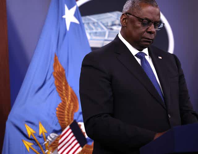 A black man with gray hair stands in front of the American flag.