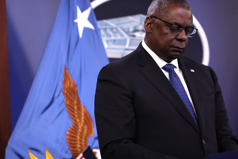 A black man with gray hair stands in front of the American flag.