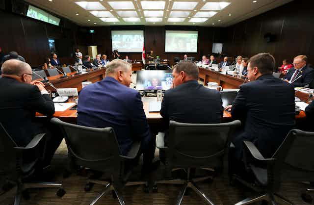 Four men are seen from the back in a committee meeting room with screens on the wall in front of them.