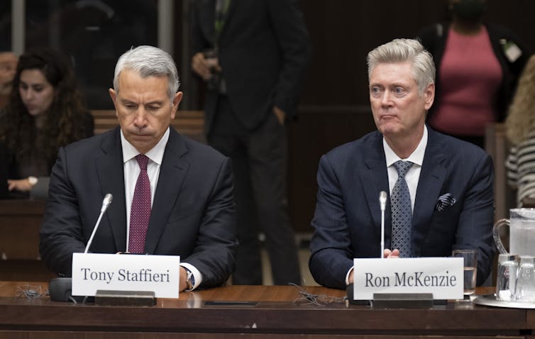 Two men sit at a table in a hearing room with microphones in front of them.