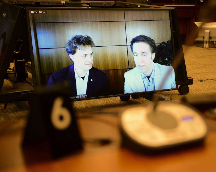 Two men are seen on a screen in a darkened room.