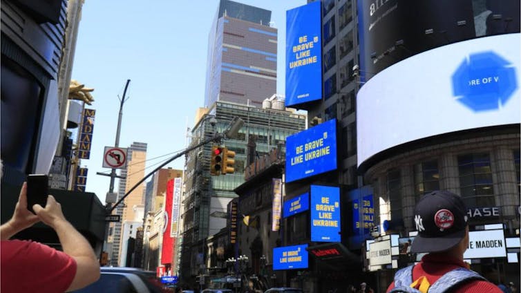 People's hands are shown holding phones and cameras, pointed at three blue billboards that say in yellow 'be brave like Ukraine